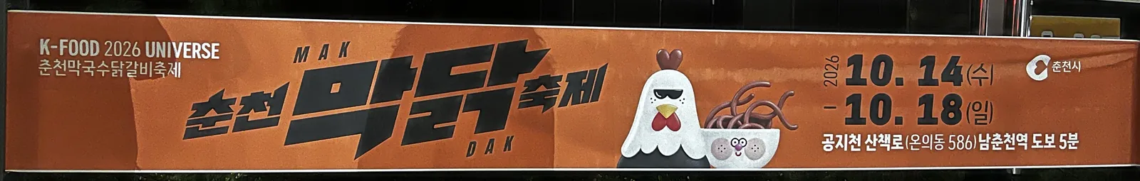춘천 길가에 걸린 2026 춘천막국수닭갈비축제 현수막 — “춘천 막닭축제”, 선글라스를 쓴 닭과 막국수 그릇 캐릭터, “2026 10. 14(수) – 10. 18(일)”, “공지천 산책로(온의동 586) 남춘천역 도보 5분”, 춘천시 로고