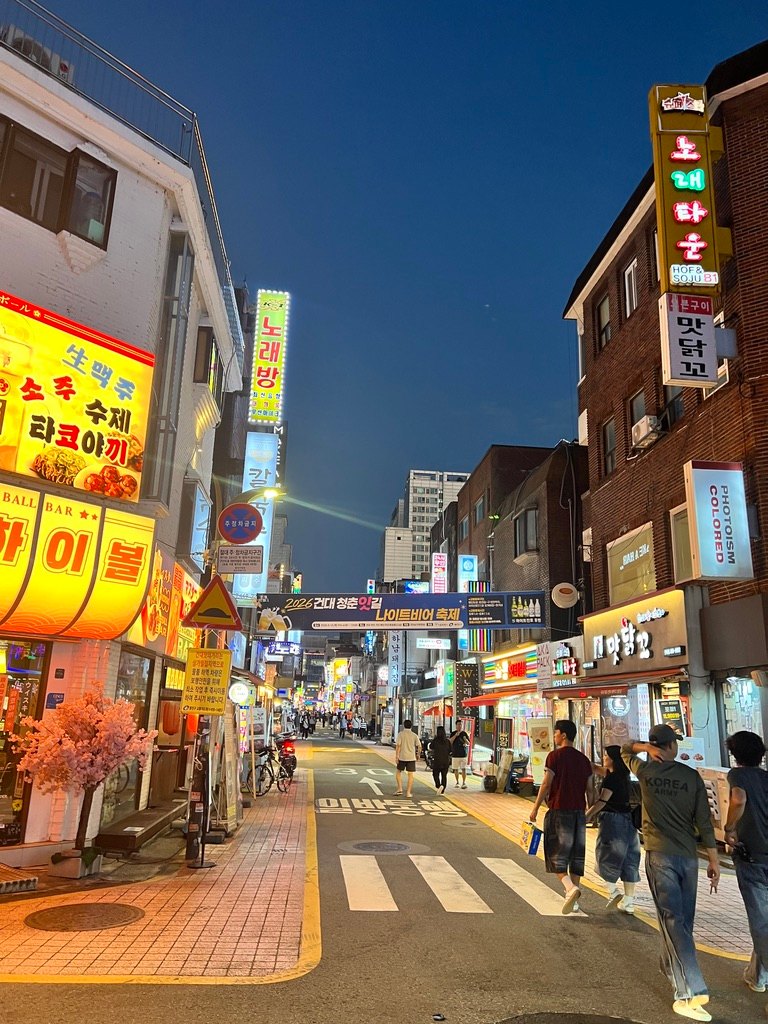 해질 무렵의 건대 맛의거리 — 「2026 건대 청춘맛길 나이트비어 축제」 현수막과 노래방·하이볼 바·닭꼬치집 간판, 일방통행 골목 (2026년 9월 초 직접 촬영)